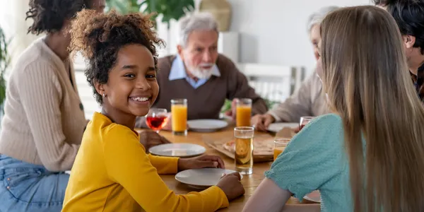 A joyful girl in a yellow shirt smiles at a dinner table with people enjoying a meal and drinks.